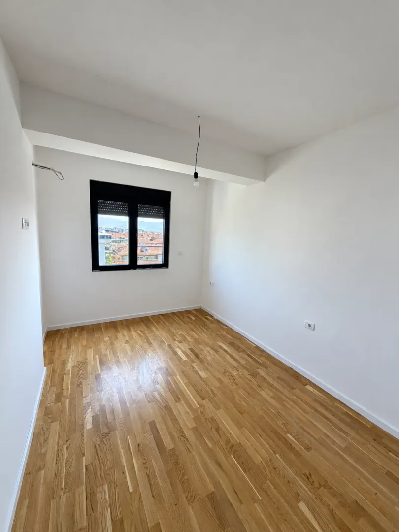 Prazna soba sa bijelim zidovima, parketom i prozorom.