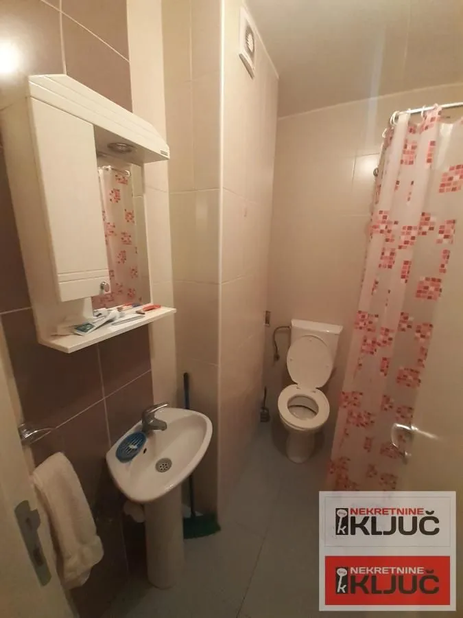 Kupatilo opremljeno umivaonikom, ogledalom i WC šoljom sa tuš zavesom.