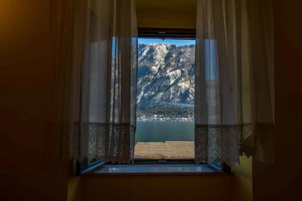 Pogled na planine i more kroz otvoren prozor sa zavjesama.