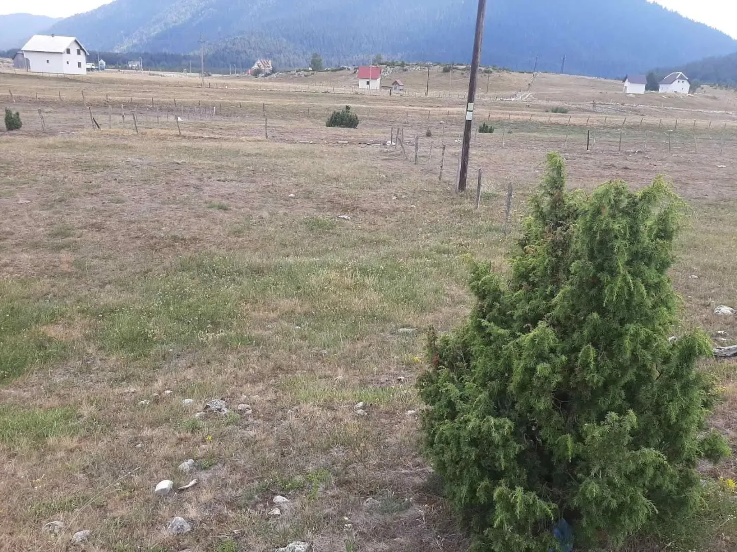 Prostrani travnati plac na planini okružen prirodom i rijetkim kućama.