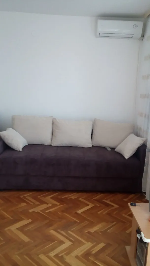 Dnevna soba sa tamnom garniturom, parketom i klima uređajem na zidu.
