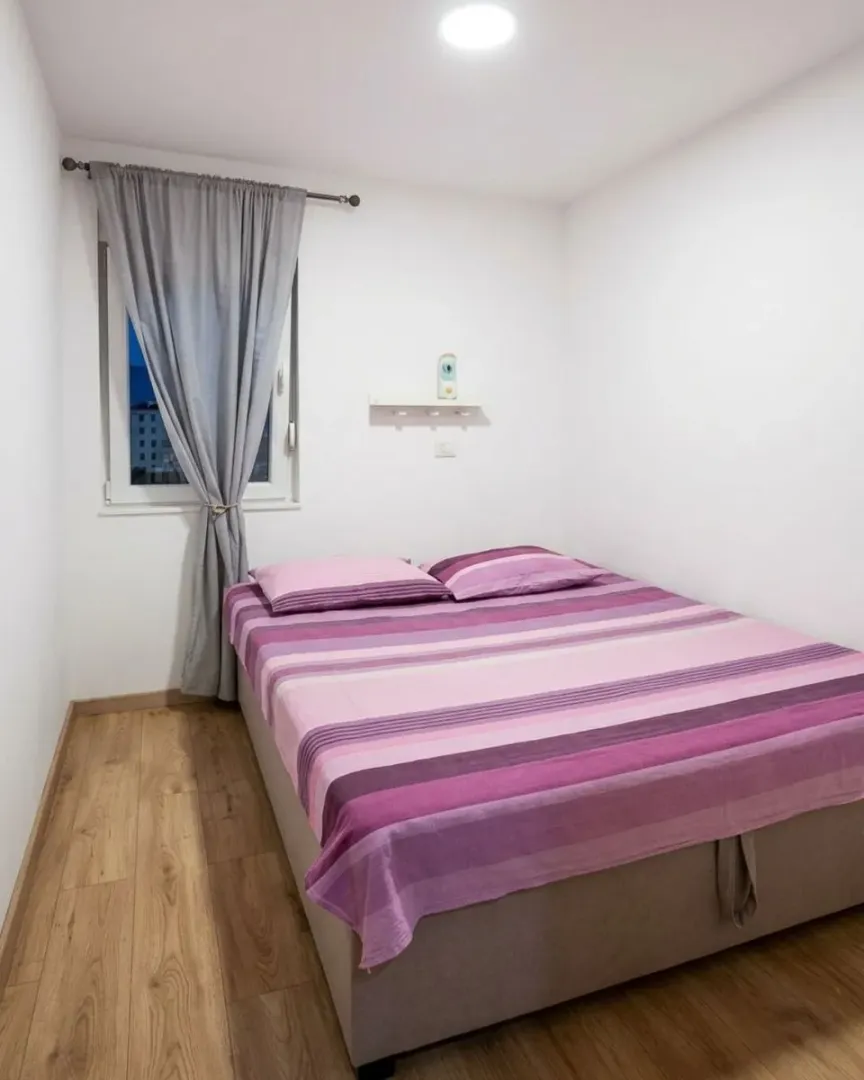Spavaća soba sa bračnim krevetom, drvenim podom i prozorom sa sivim zavjesama.