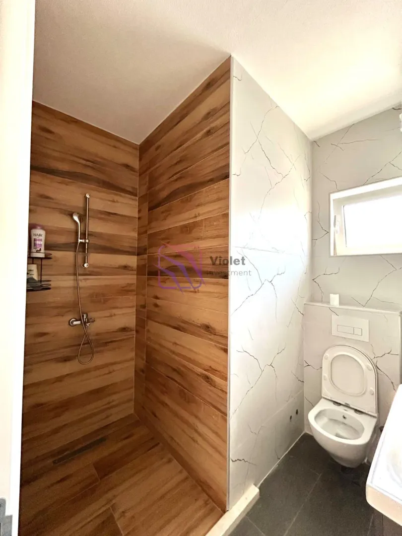 Kupatilo sa tuš kabinom obloženom pločicama u imitaciji drveta i bijelim sanitarijama.