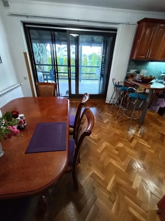 Trpezarija sa drvenim stolom i stolicama, sa izlazom na terasu i delom kuhinje u pozadini.