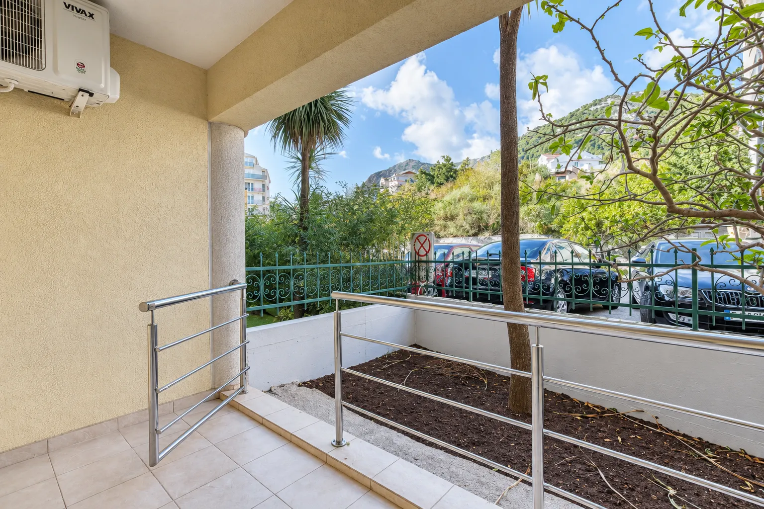 Covered terrace with a metal railing and a view of greenery and parked vehicles.