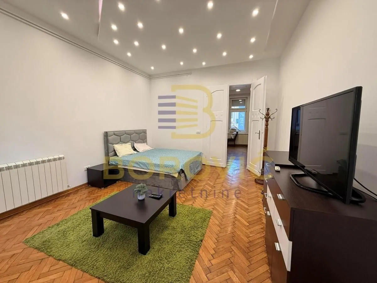 Nameštena spavaća soba sa bračnim krevetom, TV komodom i parketom na podu.