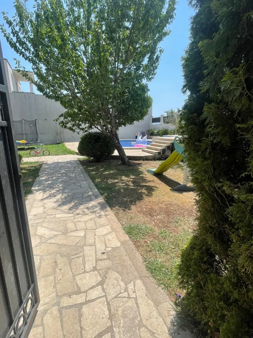 House yard with a paved walkway, lawn, trees, and children's playground equipment.