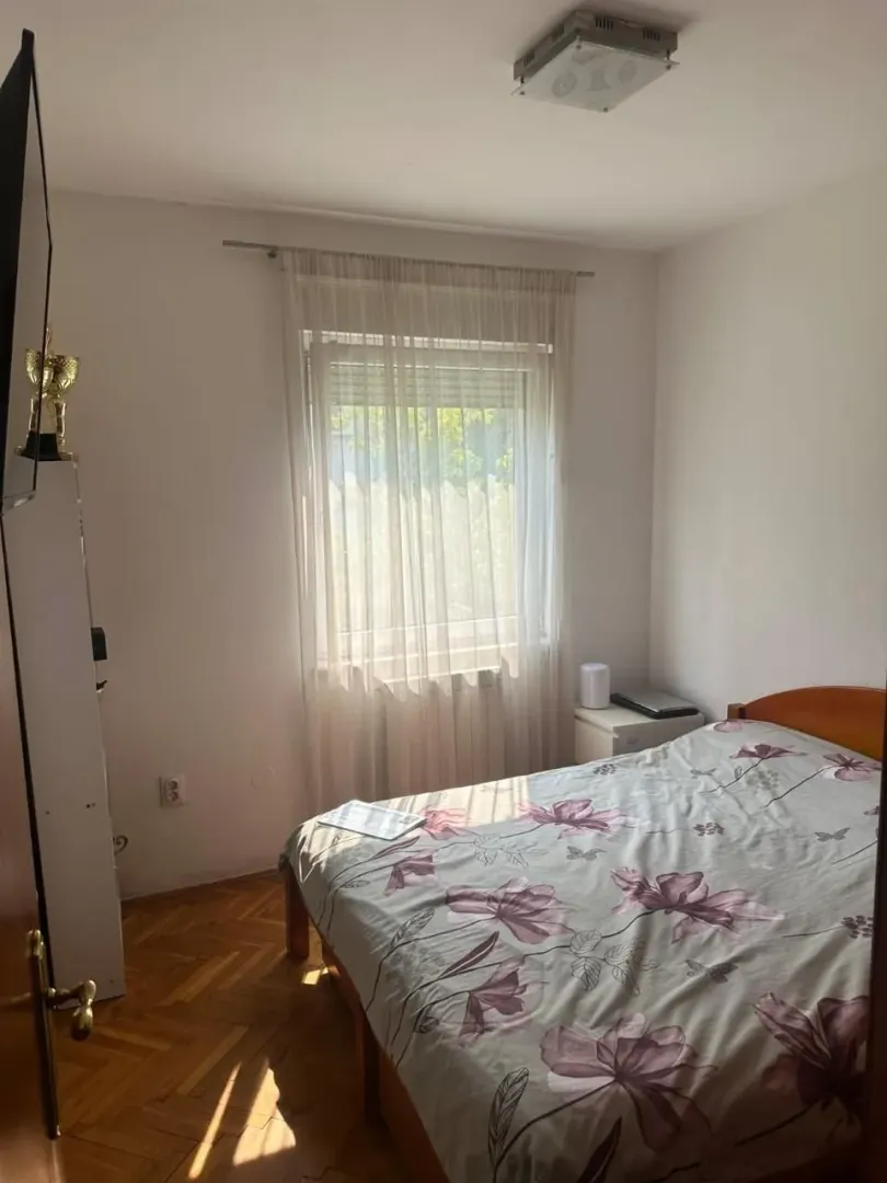 Manja spavaća soba sa bračnim krevetom, parketom i prozorom sa bijelom zavjesom.