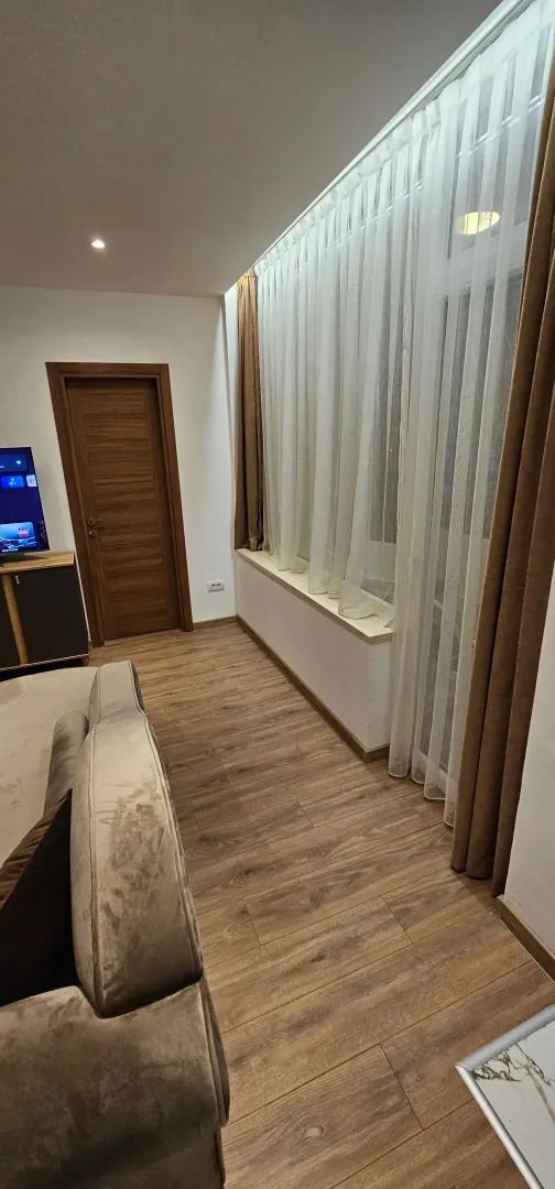 Dnevna soba sa garniturom, drvenim podom i velikim prozorom sa zavjesama.