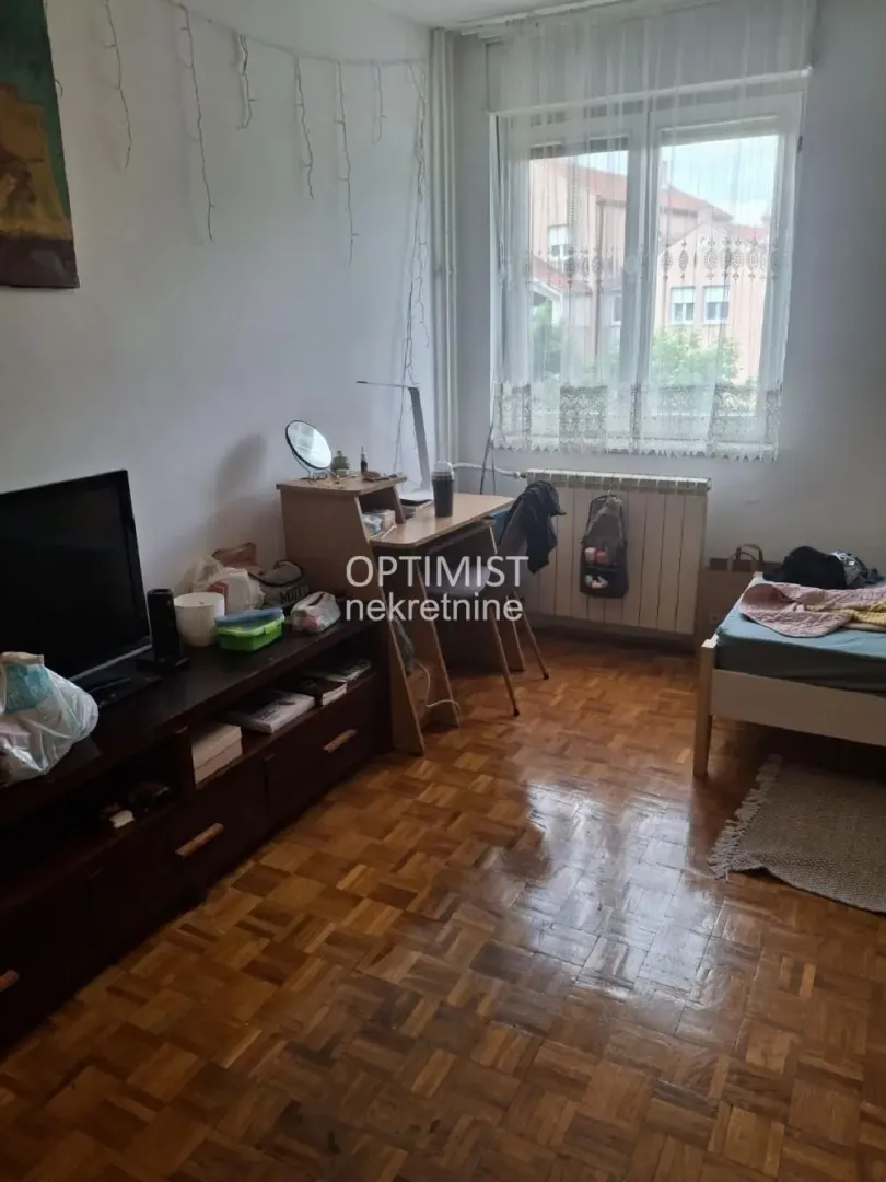 Nameštena spavaća soba sa krevetom, radnim stolom, televizorom i parketom na podu.