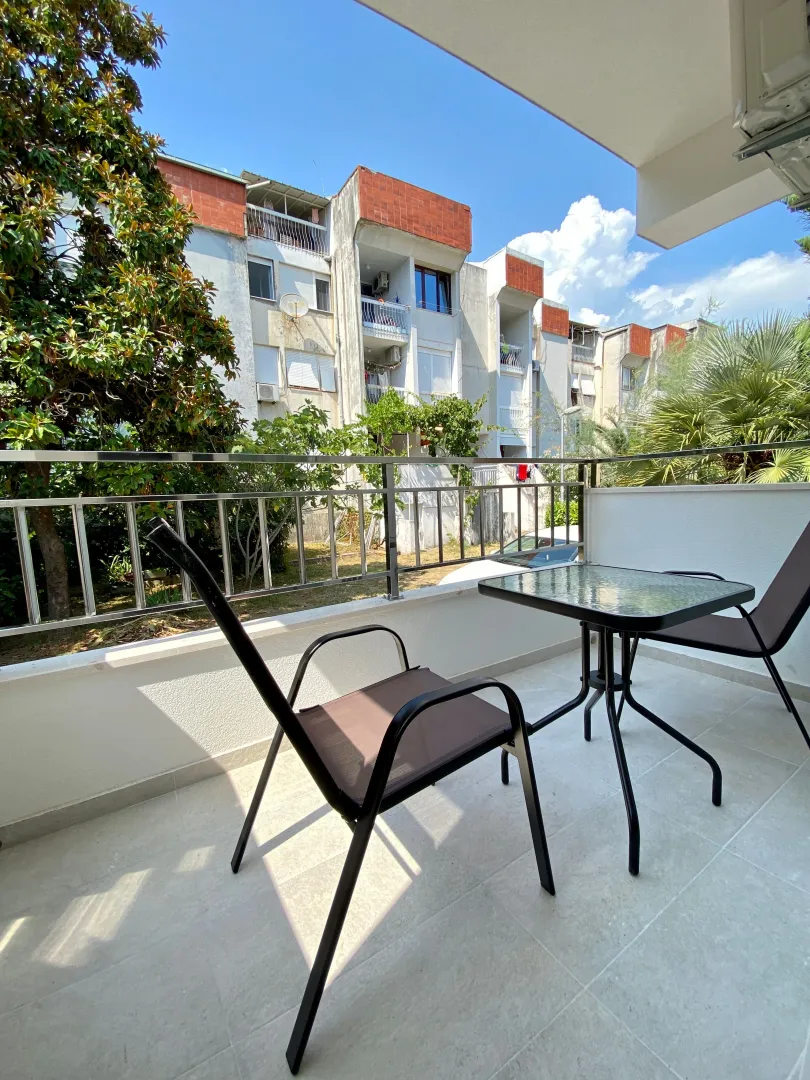 Furnished balcony with a table and chairs, overlooking greenery and a neighboring building.