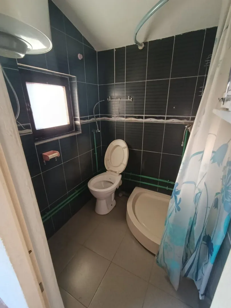 Manje kupatilo sa tamnim pločicama, wc šoljom i tuš kabinom.