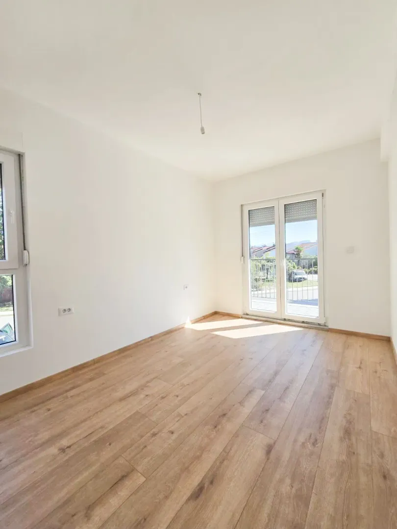 Prazna svijetla soba sa drvenim podom, bijelim zidovima i balkonskim vratima.