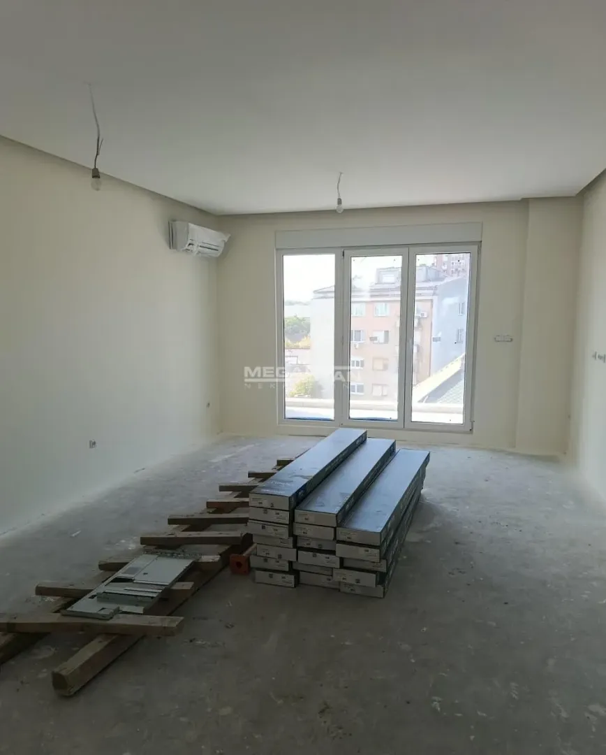 Prazna neuuređena prostorija sa paketom spakovanim na podu i balkonskim vratima.