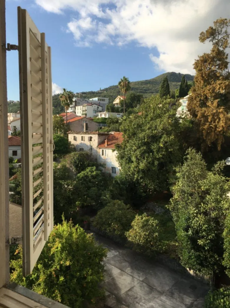 Pogled sa prozora na okolno zelenilo, kuće i brda u daljini.