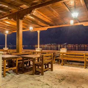 Natkrivena terasa sa drvenim stolovima i klupama sa pogledom na more u noćnim satima.