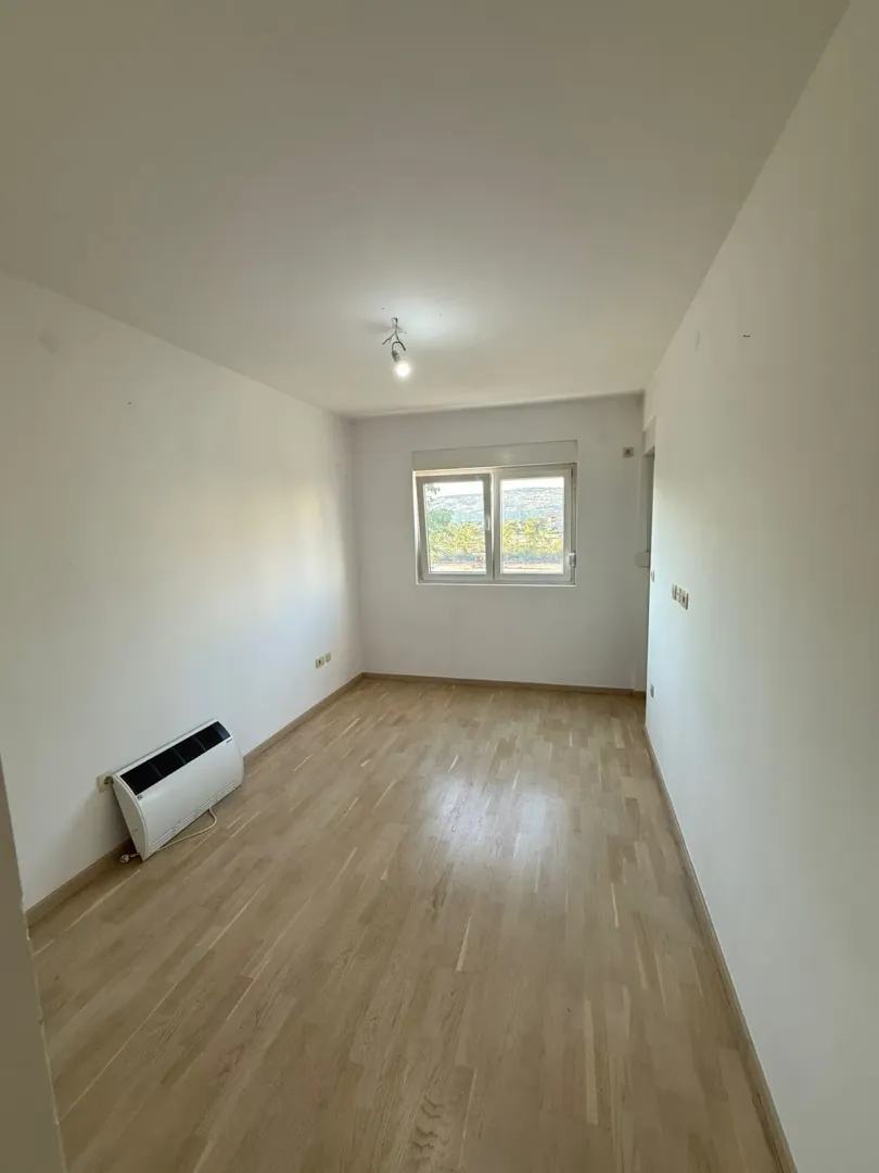Prazna soba sa bijelim zidovima, drvenim podom i prozorom.