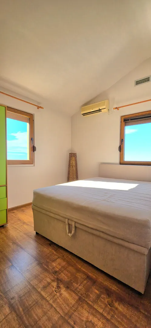 Bright bedroom with a double bed, wooden floor, and two windows.