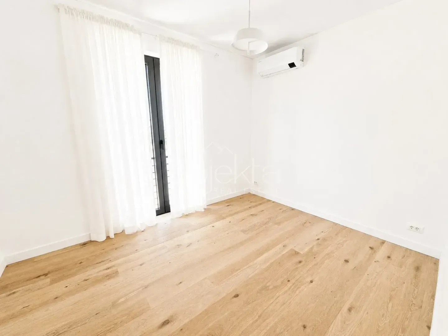 Empty bright room with wooden flooring, an air conditioning unit, and balcony doors with white curtains.