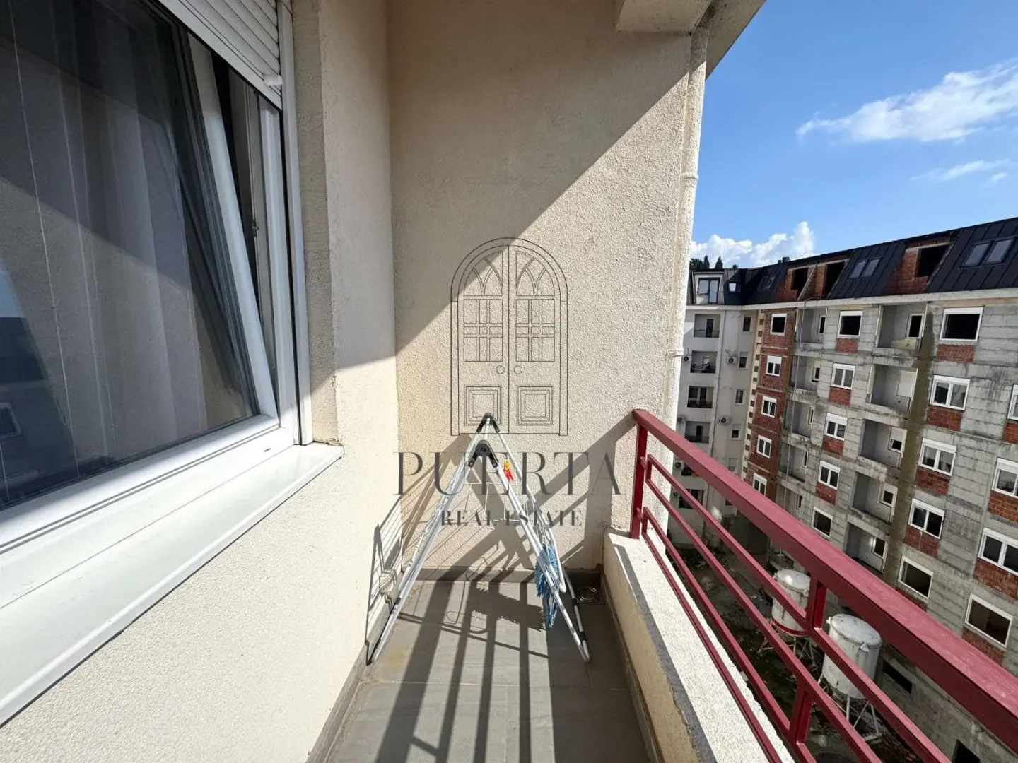 Manji uski balkon sa crvenom ogradom i pogledom na zgradu u izgradnji.