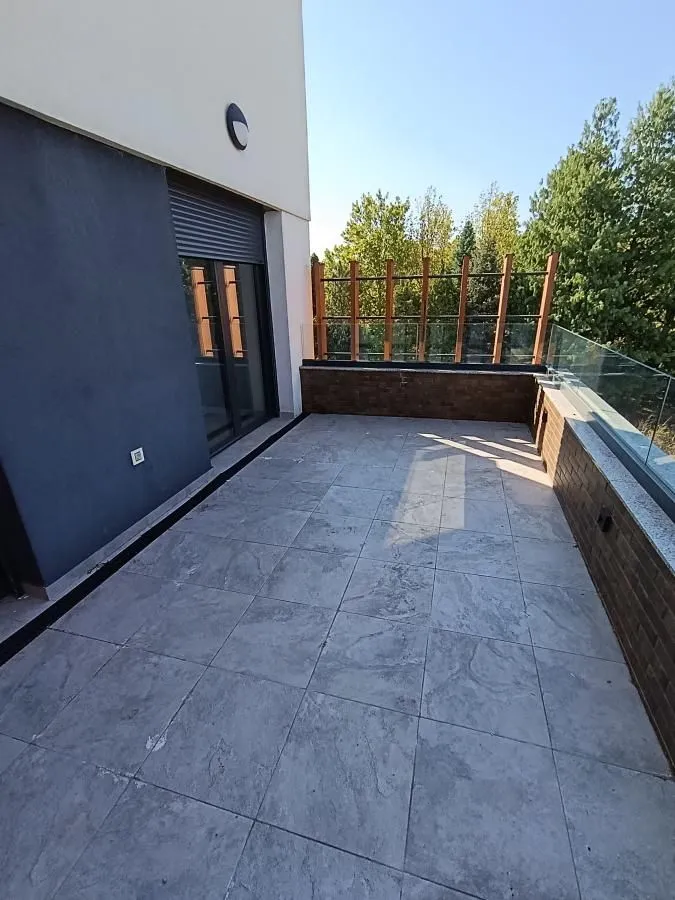 Spacious tiled terrace with a glass railing and a view of greenery.