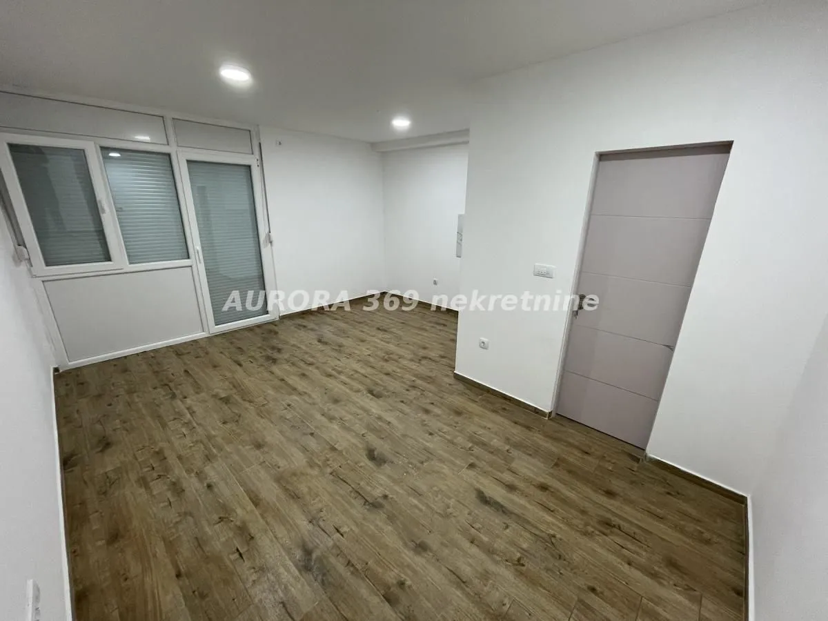 Prazna soba sa drvenim podom, belim zidovima i vratima koja vode na balkon.