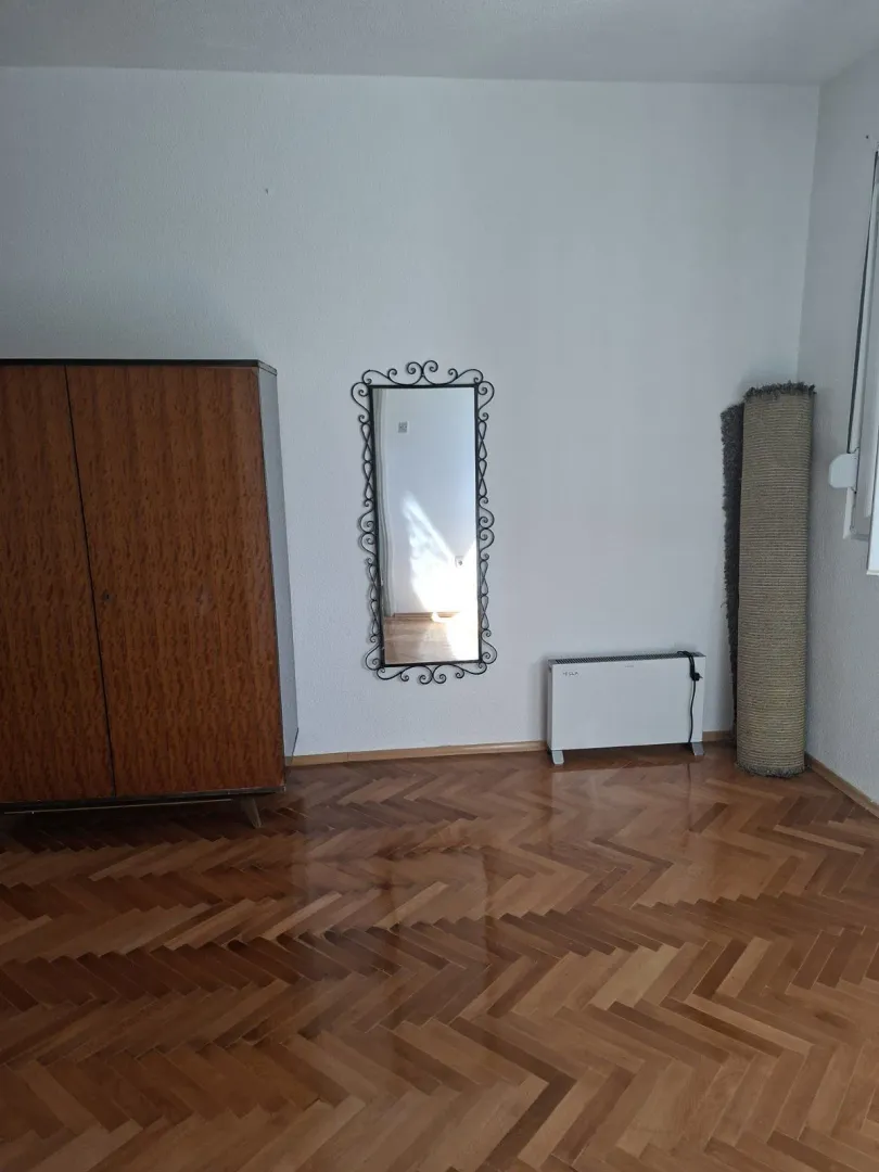 Polunamještena soba sa drvenim ormarom, ogledalom i parketom.
