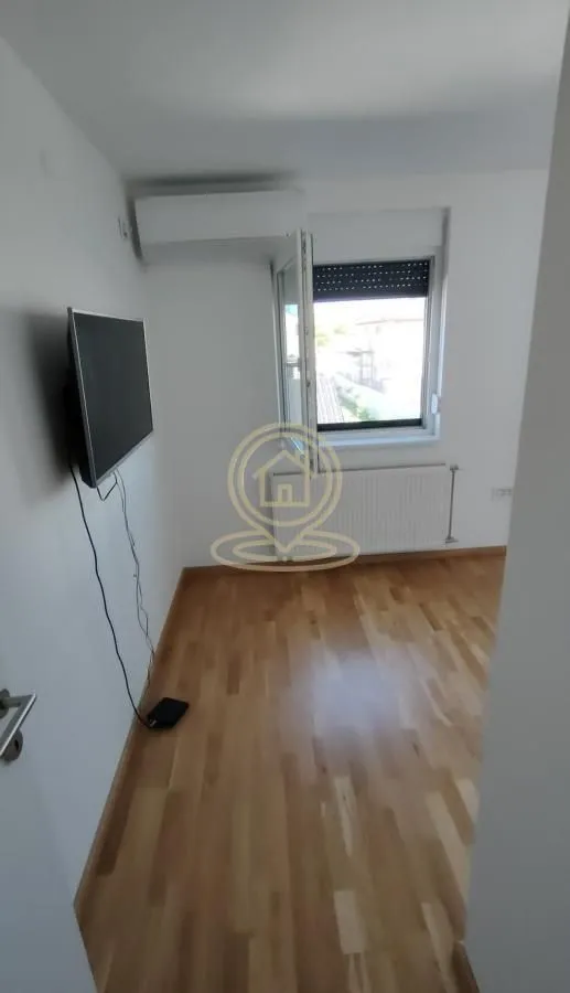 Prazna spavaća soba sa drvenim podom, televizorom na zidu i prozorom.