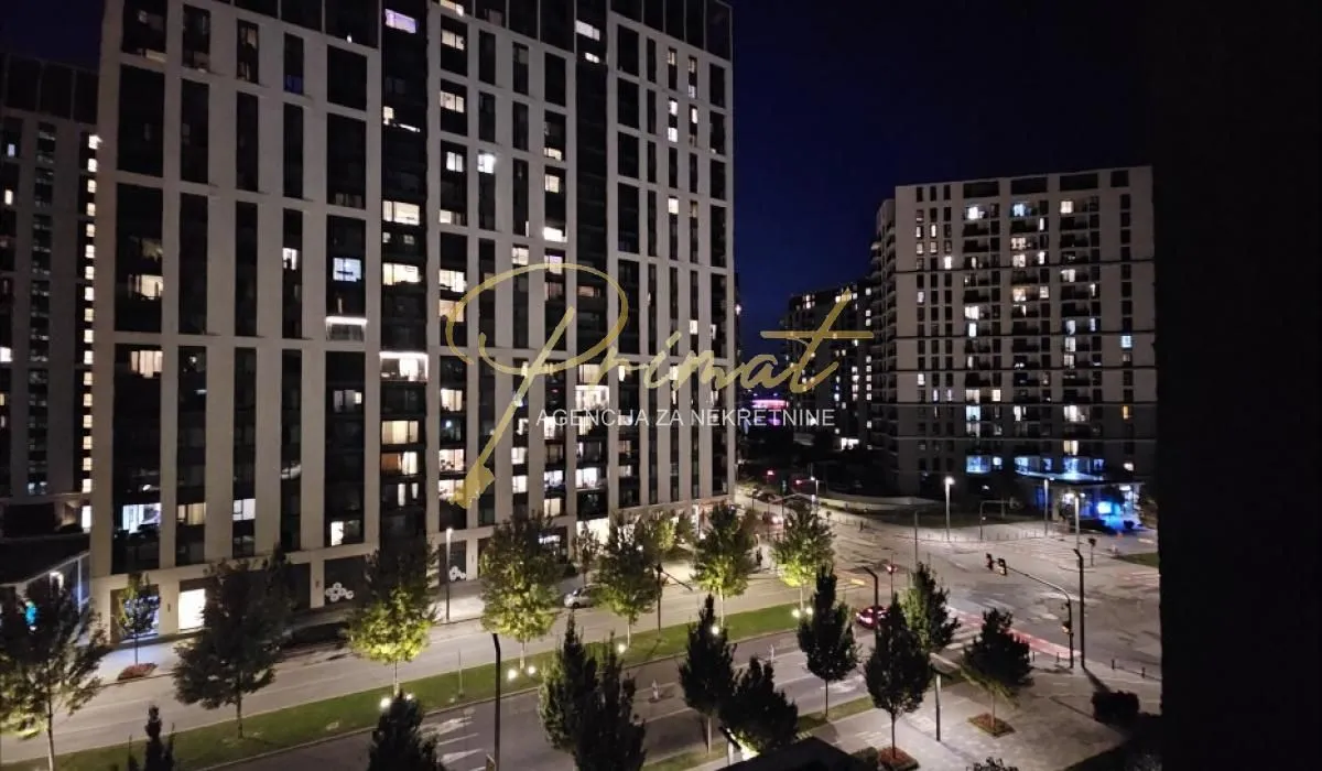 Noćni pogled sa prozora na osvetljene gradske ulice i moderne stambene zgrade.