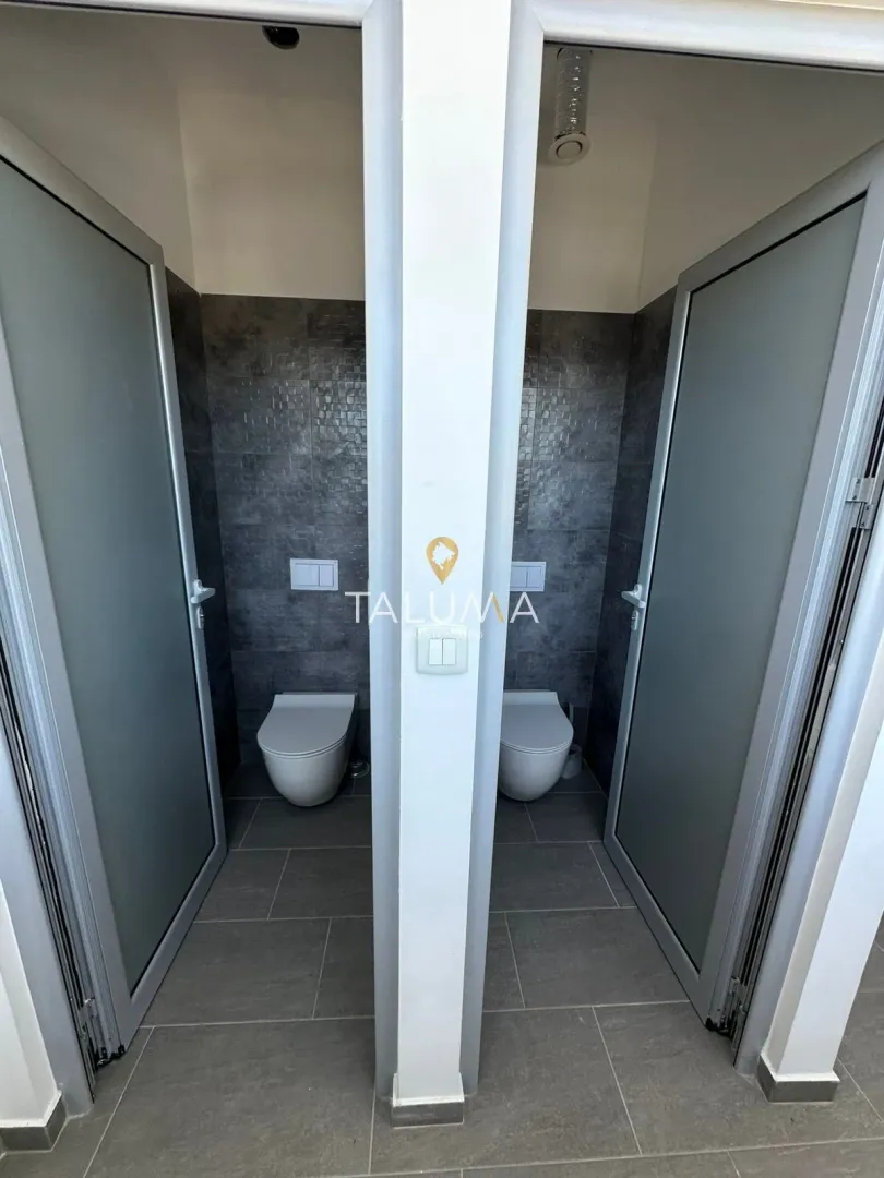 View of two separate toilets with dark tiles and glass doors.