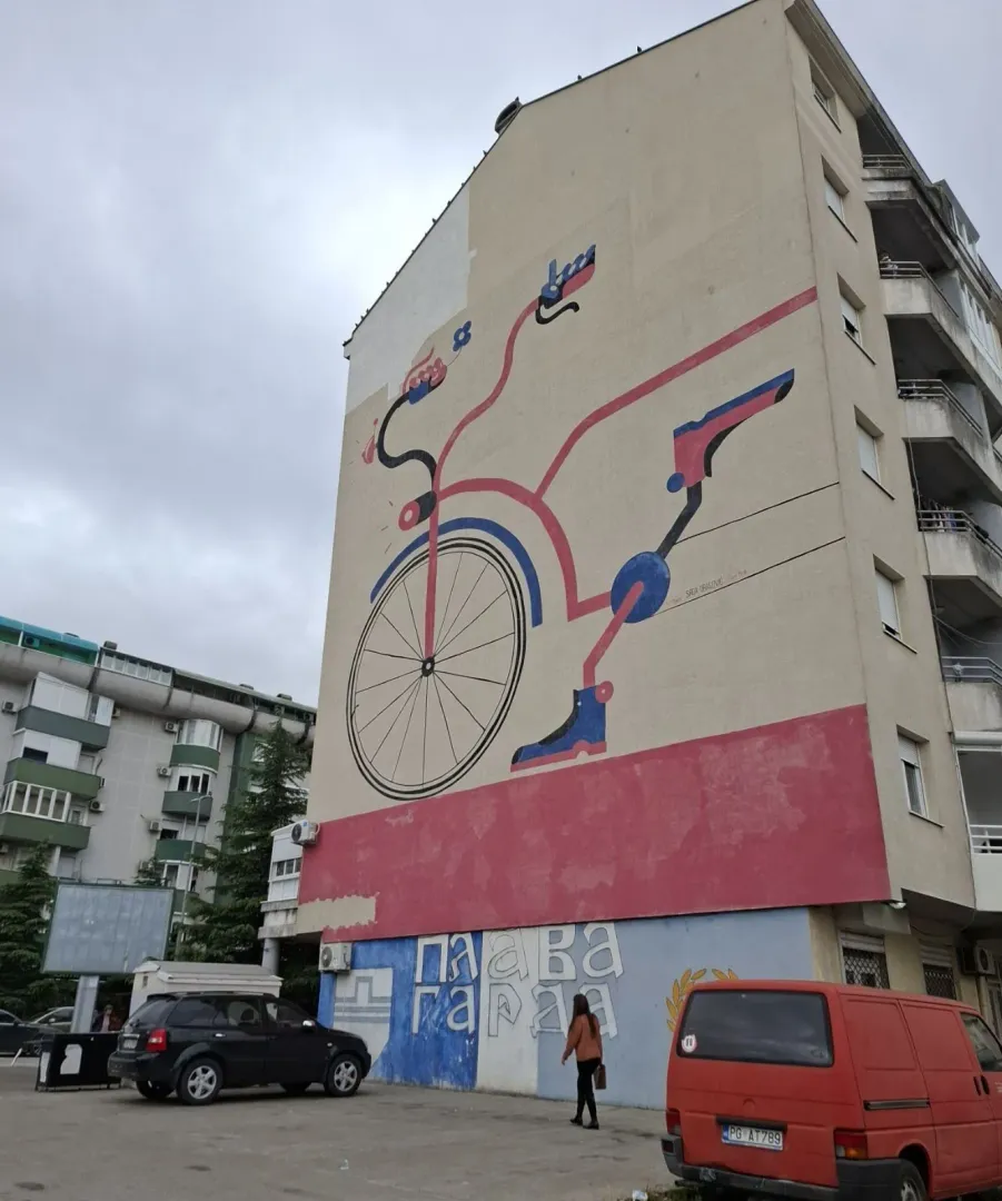 Vanjski izgled stambene zgrade sa velikim muralom na fasadi i parkingom ispred.