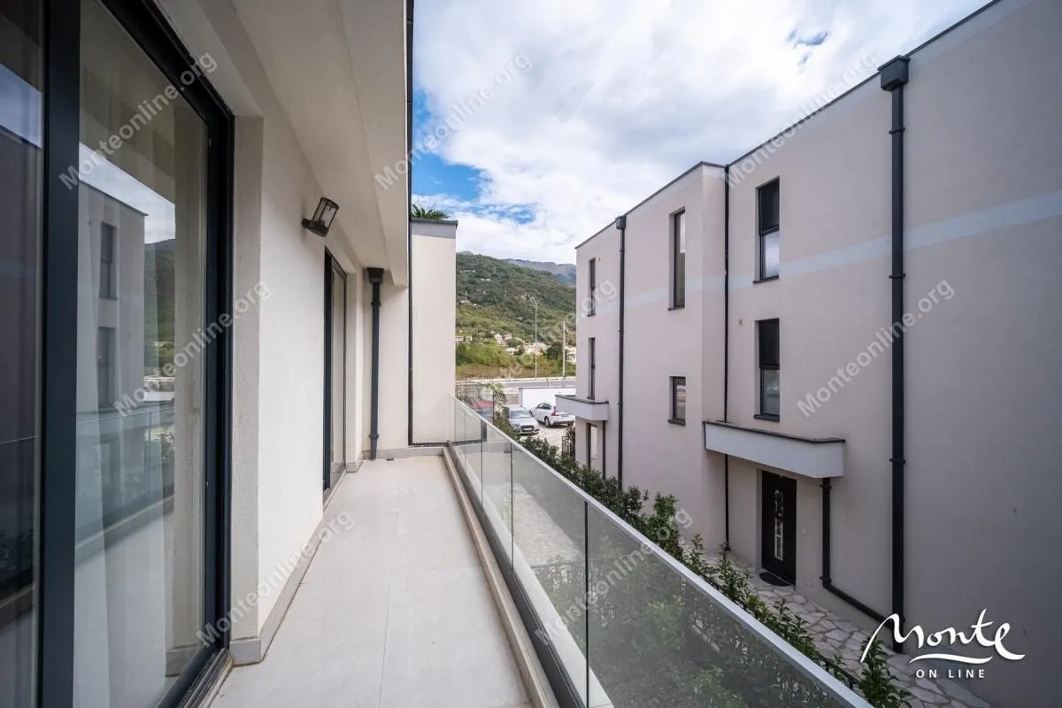 Dugačak popločani balkon sa staklenom ogradom i pogledom na okolne zgrade i brda.