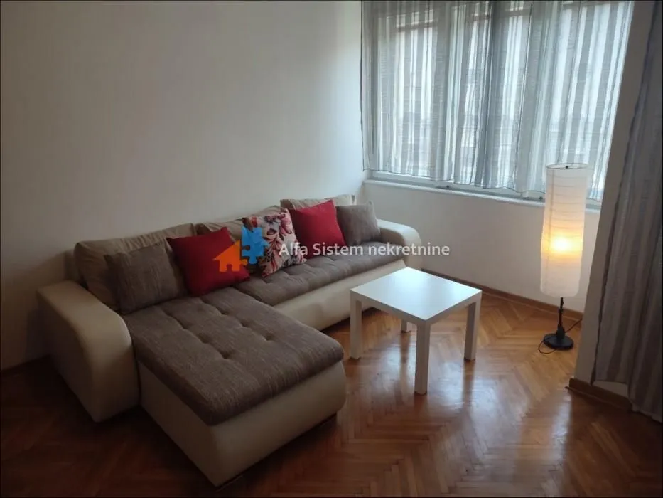 Dnevna soba sa ugaonom garniturom, stočićem i lampom pored prozora.