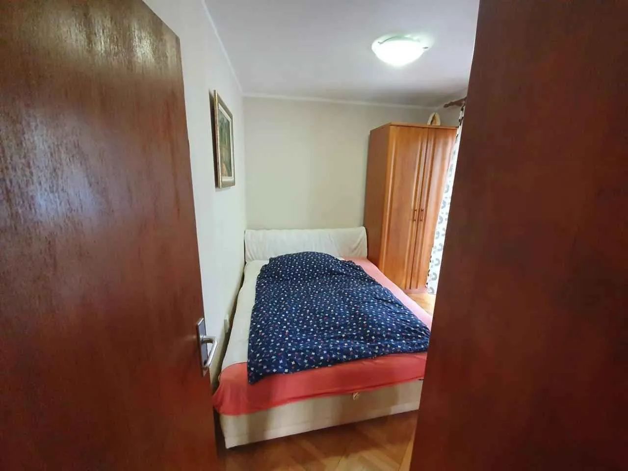 Pogled na spavaću sobu sa krevetom i drvenim ormarom kroz otvorena vrata.