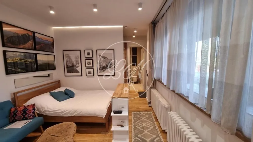 Nameštena spavaća soba sa bračnim krevetom, foteljom i velikim prozorima.