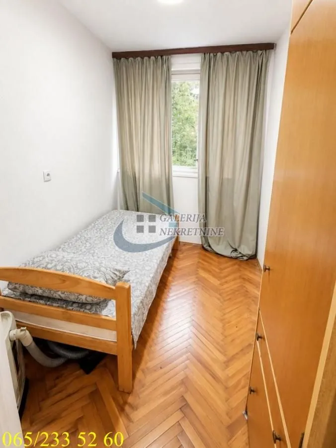 Manja spavaća soba sa krevetom, drvenim podom i prozorom sa zavesama.