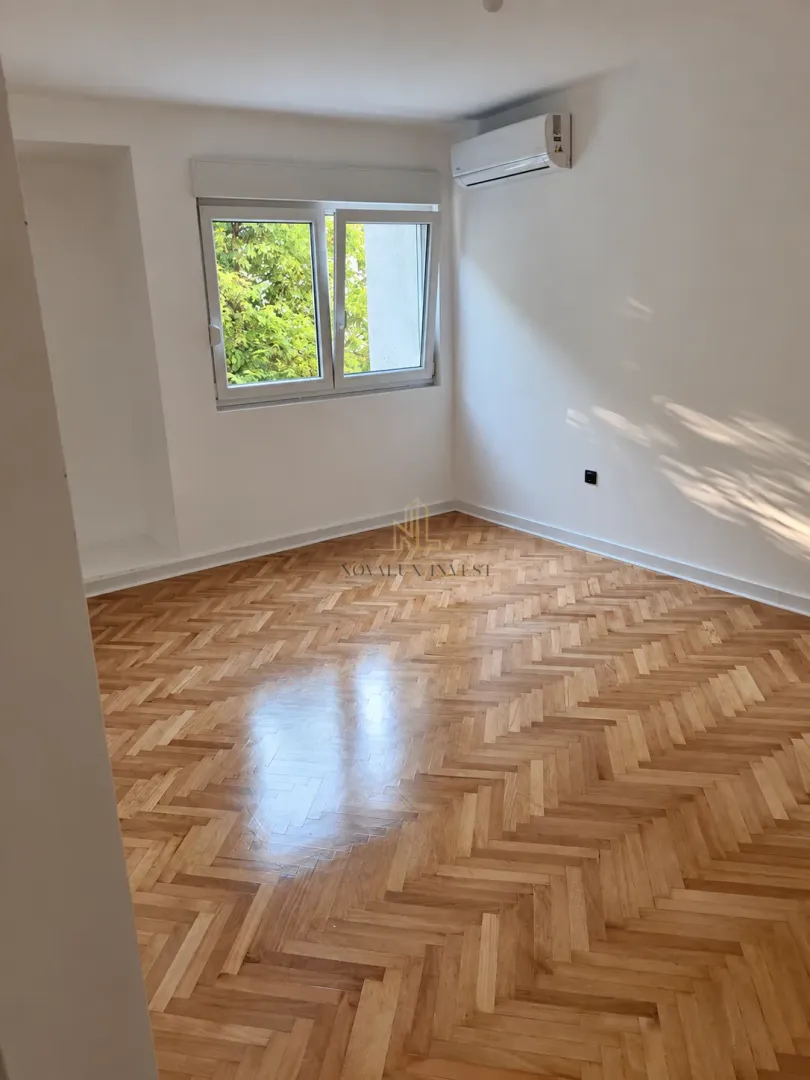 Prazna soba sa drvenim parketom, prozorom i klima uređajem.
