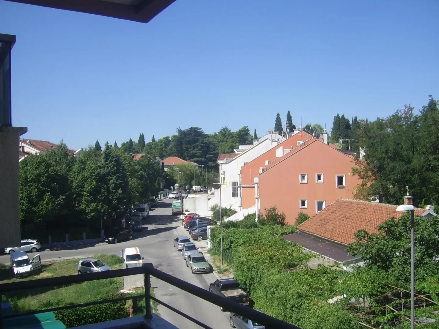 Pogled sa balkona na ulicu i okolne zgrade pod vedrim nebom.