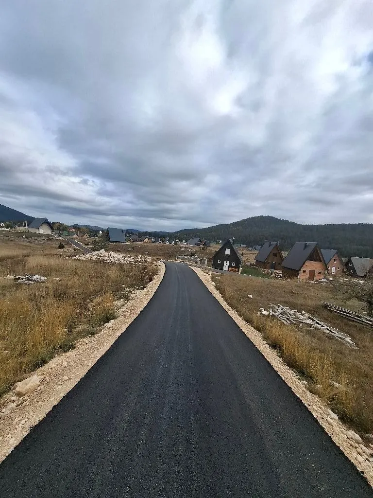 Asfaltni put koji vodi kroz naselje sa vikendicama i brdovitim pejzažem u pozadini.