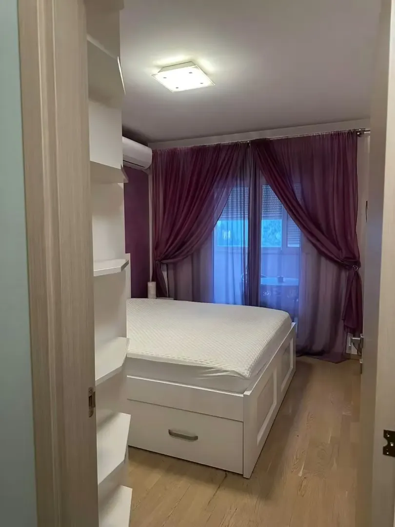 Spavaća soba sa bračnim krevetom, bijelim elementima i ljubičastim zavesama.