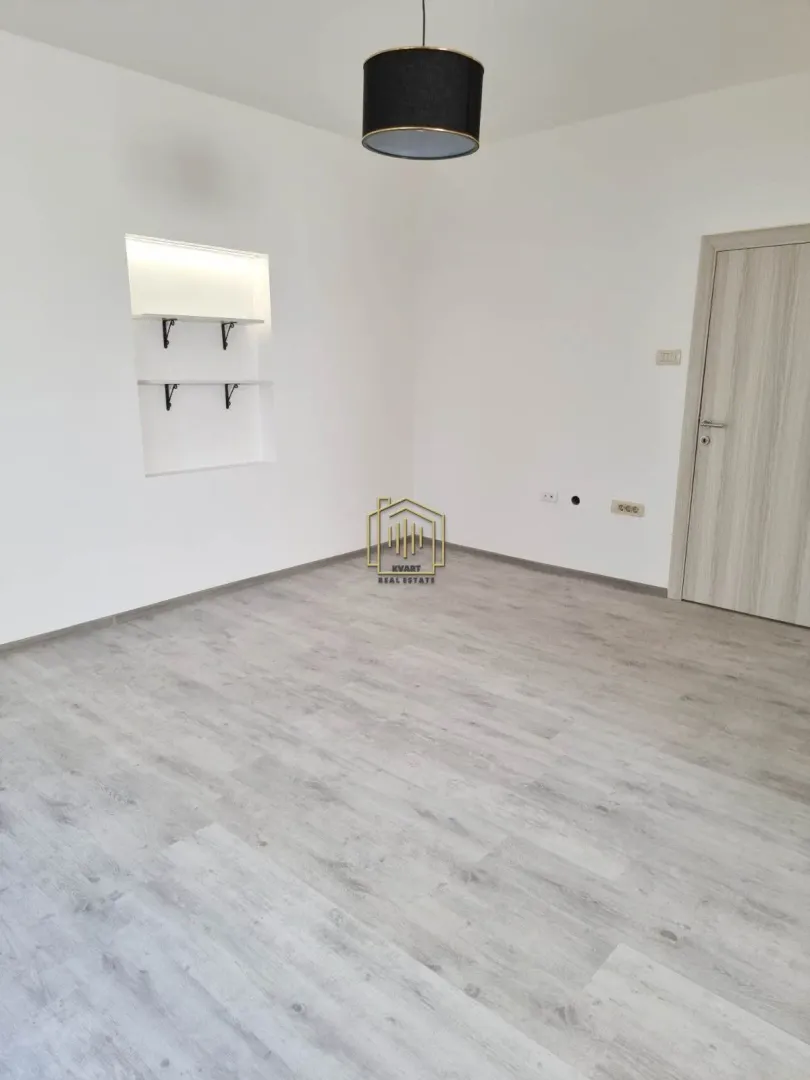 Empty room with white walls, wooden flooring, a door, and a ceiling light.