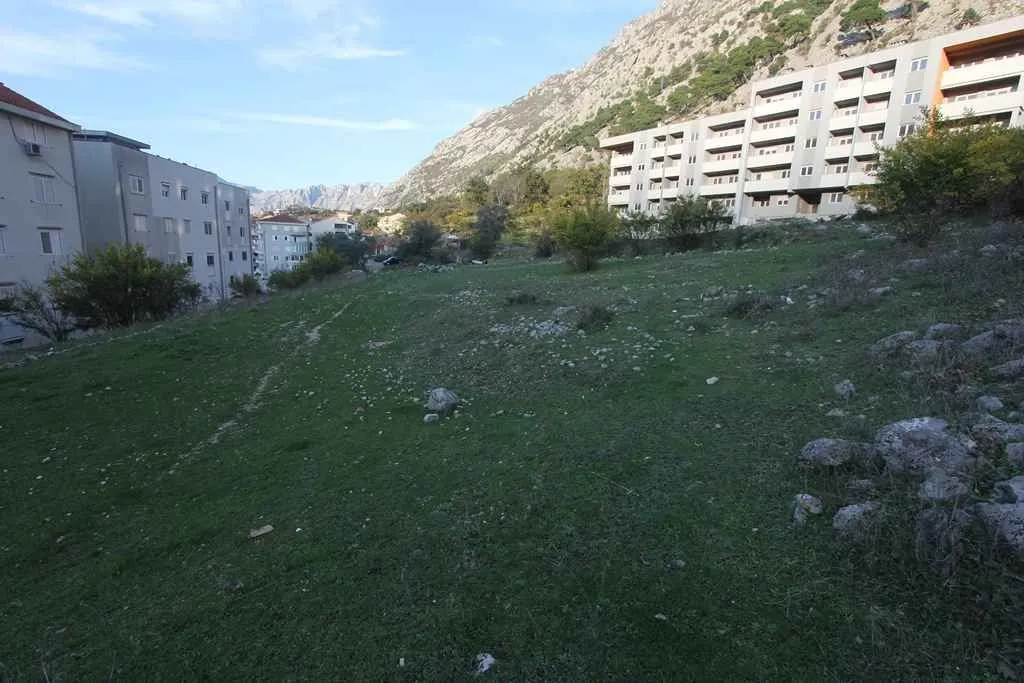 Travnati plac za gradnju okružen stambenim zgradama i brdima u pozadini.