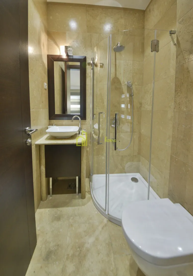Kupatilo obloženo bež pločicama sa tuš kabinom, umivaonikom i WC šoljom.