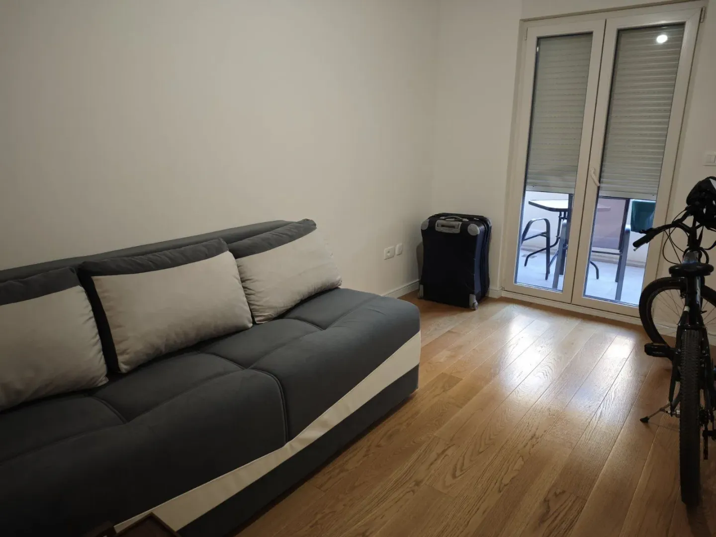Svijetla soba sa sivom garniturom, parketom i izlazom na balkon.