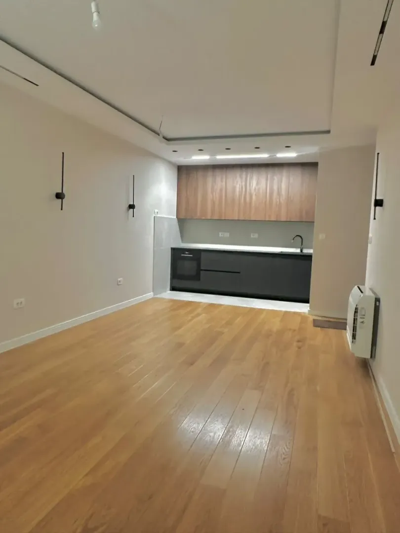 Living room with wooden flooring and a built-in kitchen in the background.