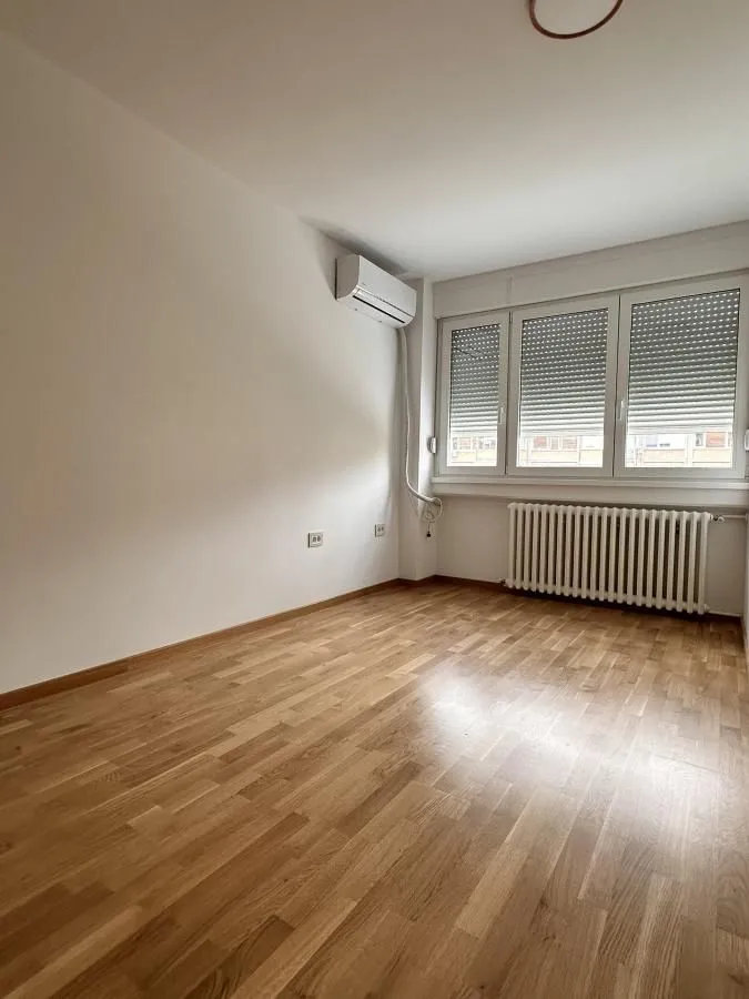 Prazna soba sa parketom, klima uređajem i prozorom sa roletnama.
