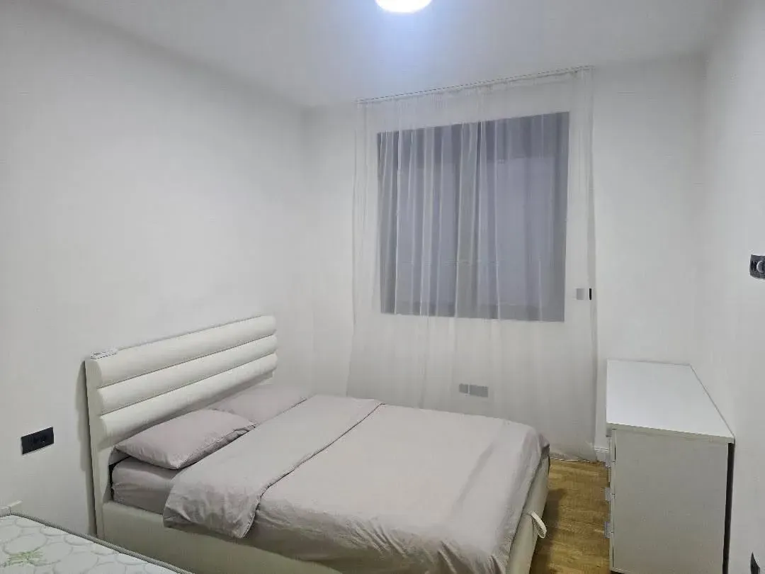 Svijetla spavaća soba sa bračnim krevetom, bijelom komodom i prozorom sa zavjesom.