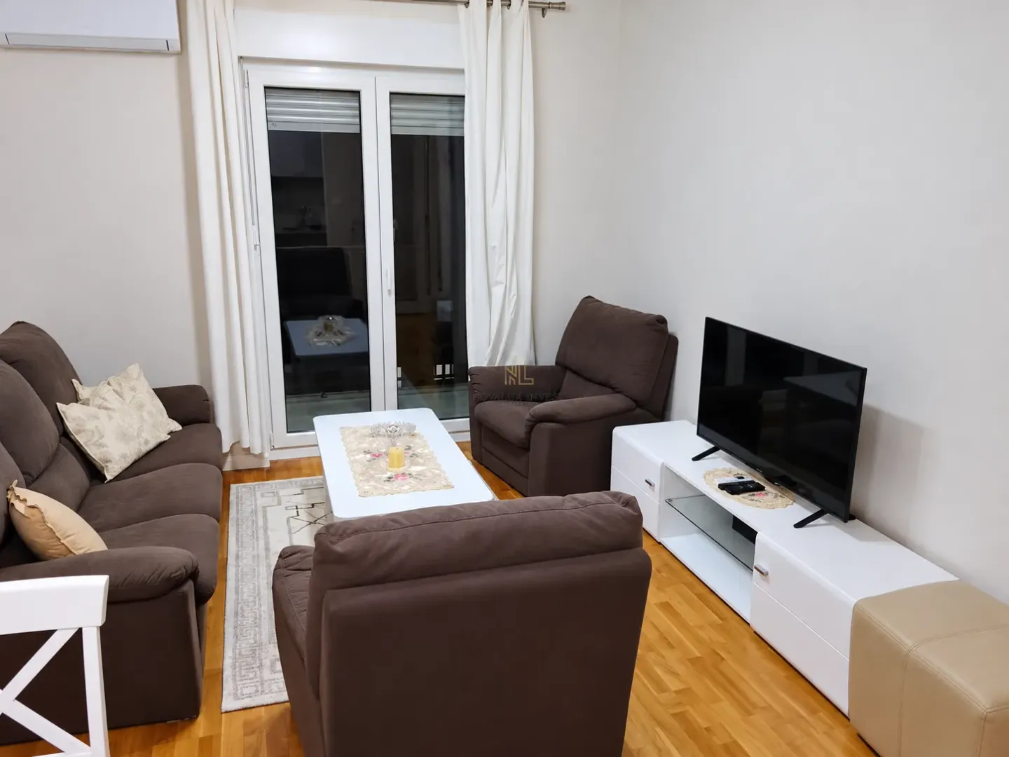 Dnevna soba sa garniturom za sjedenje, TV komodom i balkonskim vratima.