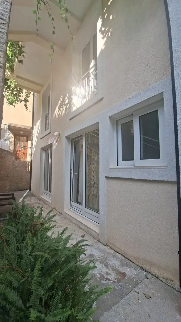Pogled na spoljašnjost kuće sa popločanim dvorištem i zelenilom.