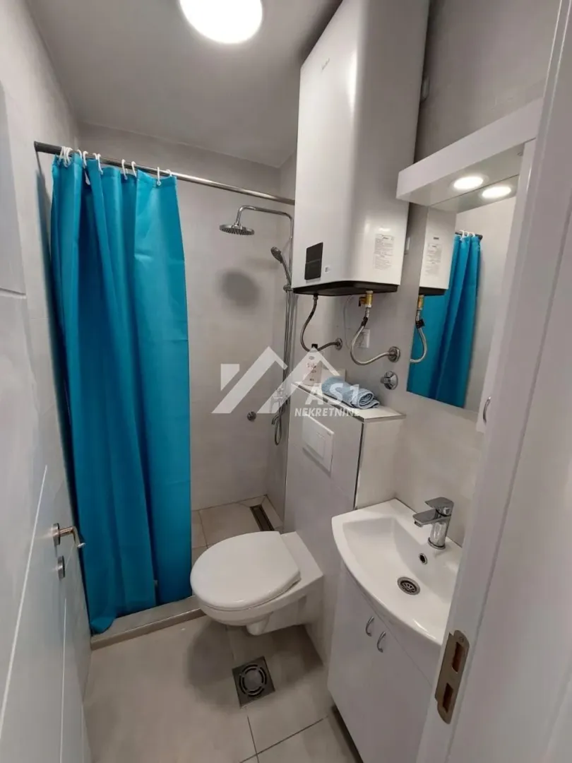Manje kupatilo sa tuš kabinom i plavom zavesom, wc šoljom i umivaonikom.